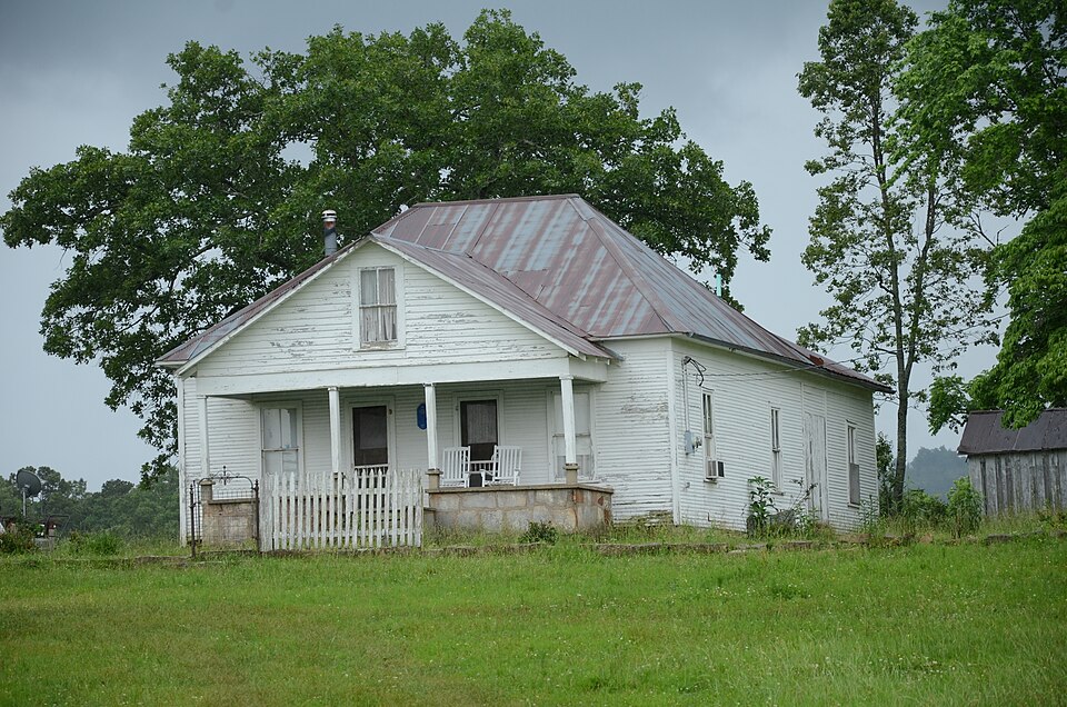 A white frame farmhouse with a tin roof, a front porch and a picket fence, under a big tree on a grey day