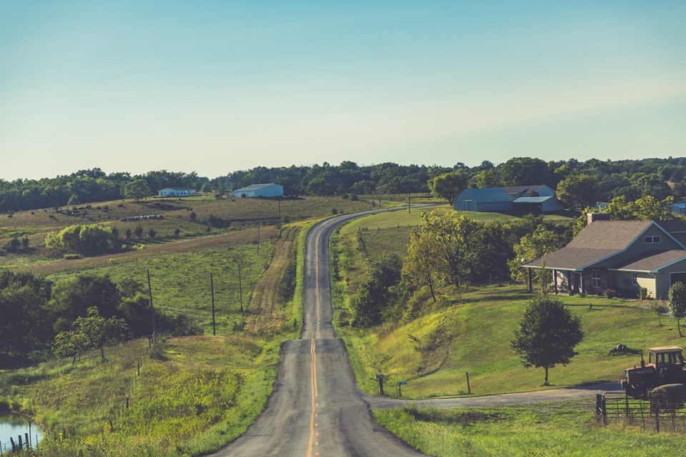 A narrow country road dipping and rising between farmhouses, sheds and hay fields toward a wooded horizon in evening light