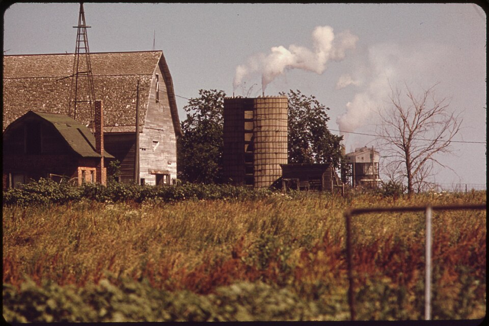 An old gambrel-roofed barn, a silo and outbuildings behind an overgrown field, with white smoke pouring from an industrial plant just beyond them