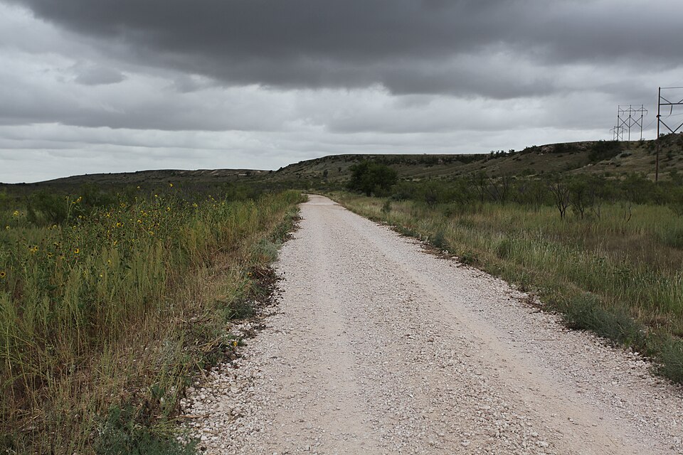 A white caliche road running through grass and sunflowers toward a low rocky escarpment under heavy grey cloud