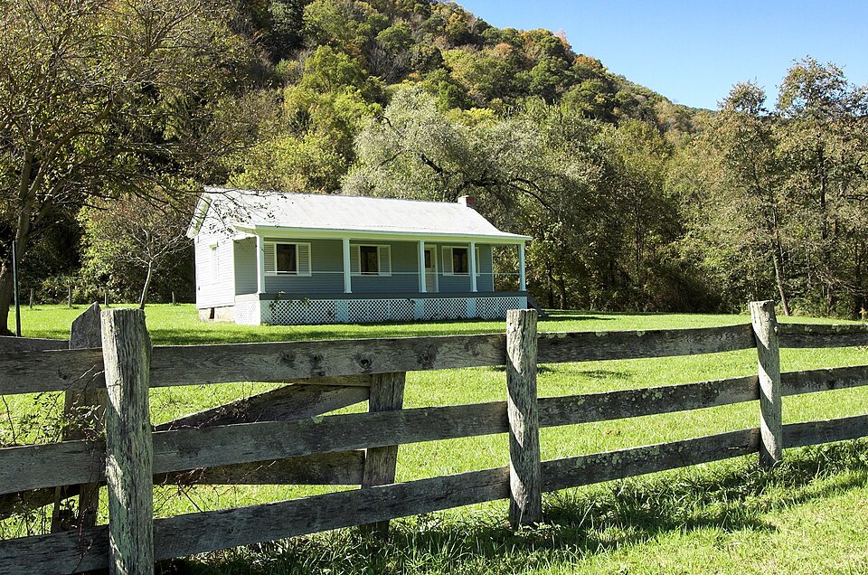 A small farmhouse with a long front porch in a mown field, seen across a split-rail fence, with a wooded hillside behind