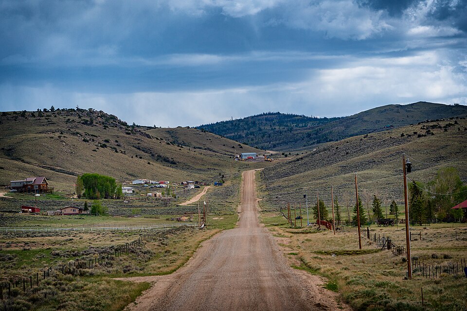 A dirt road running straight between sagebrush hills, past a scatter of houses, sheds and fenced lots, under a dark storm sky